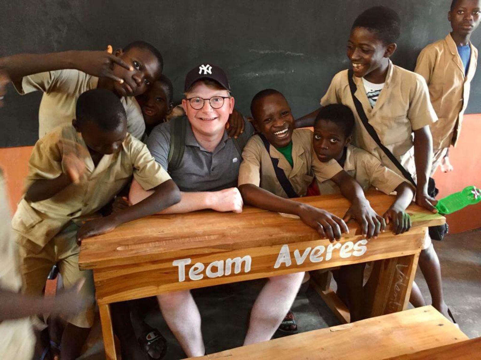 Mitarbeiter AVERES sitzt auf Schulbank mit Aufschrift Team Averes zusammen mit lachenden Schülern aus Benin mit Schuluniform