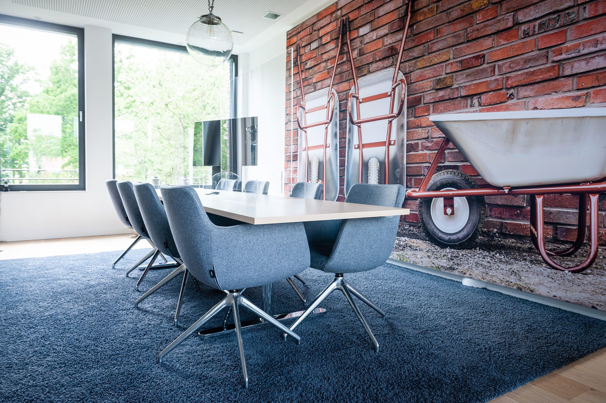 Kleiner Konferenzraum mit großen Fenstern, einem großen Tisch und gemütlichen Stühlen in blau mit blauem Teppich, im Hintergrund ein Fernseher, an der Wand ein sehr großes Bild mit Schubkarren vor Klinkermauer